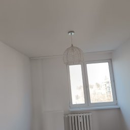 Maiki - Jasne wnętrze po malowaniu: białe ściany, okno z widokiem na miasto, grzejnik i drewniana podłoga. W centralnej części wisi ozdobna lampa.