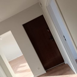 Maiki - Ciemne drzwi wejściowe z ościeżnicą, widoczne z perspektywy korytarza z jasną podłogą i białymi ścianami. Minimalistyczny design, świeżo po remoncie.