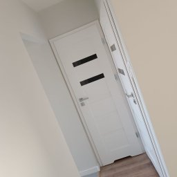 Maiki - Białe drzwi wewnętrzne z czarnymi, prostokątnymi przeszkleniami, zamontowane w jasnym korytarzu z panelami podłogowymi. Widok z dołu, lekko ukośny.