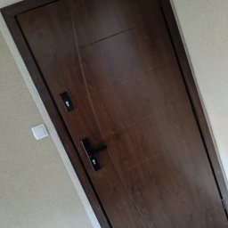 Maiki - Ciemnobrązowe, nowoczesne drzwi wewnętrzne z czarną klamką i zamkiem, zamontowane w ościeżnicy. Widoczny fragment ściany z włącznikiem światła.