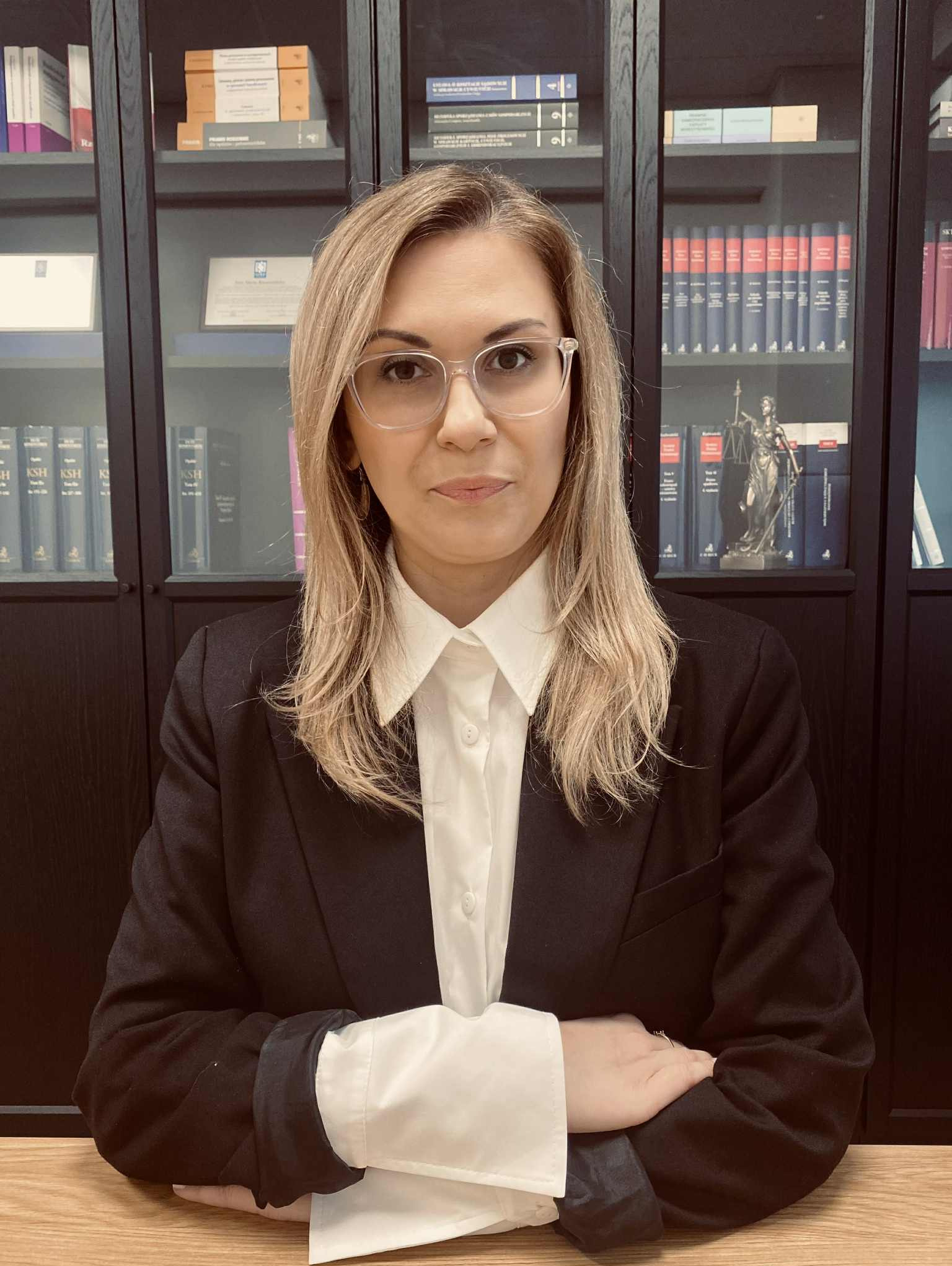 Portret kobiety w okularach, ubrana w czarną marynarkę i białą koszulę, na tle regału z książkami i figurką Temidy. Profesjonalny wizerunek prawnika.