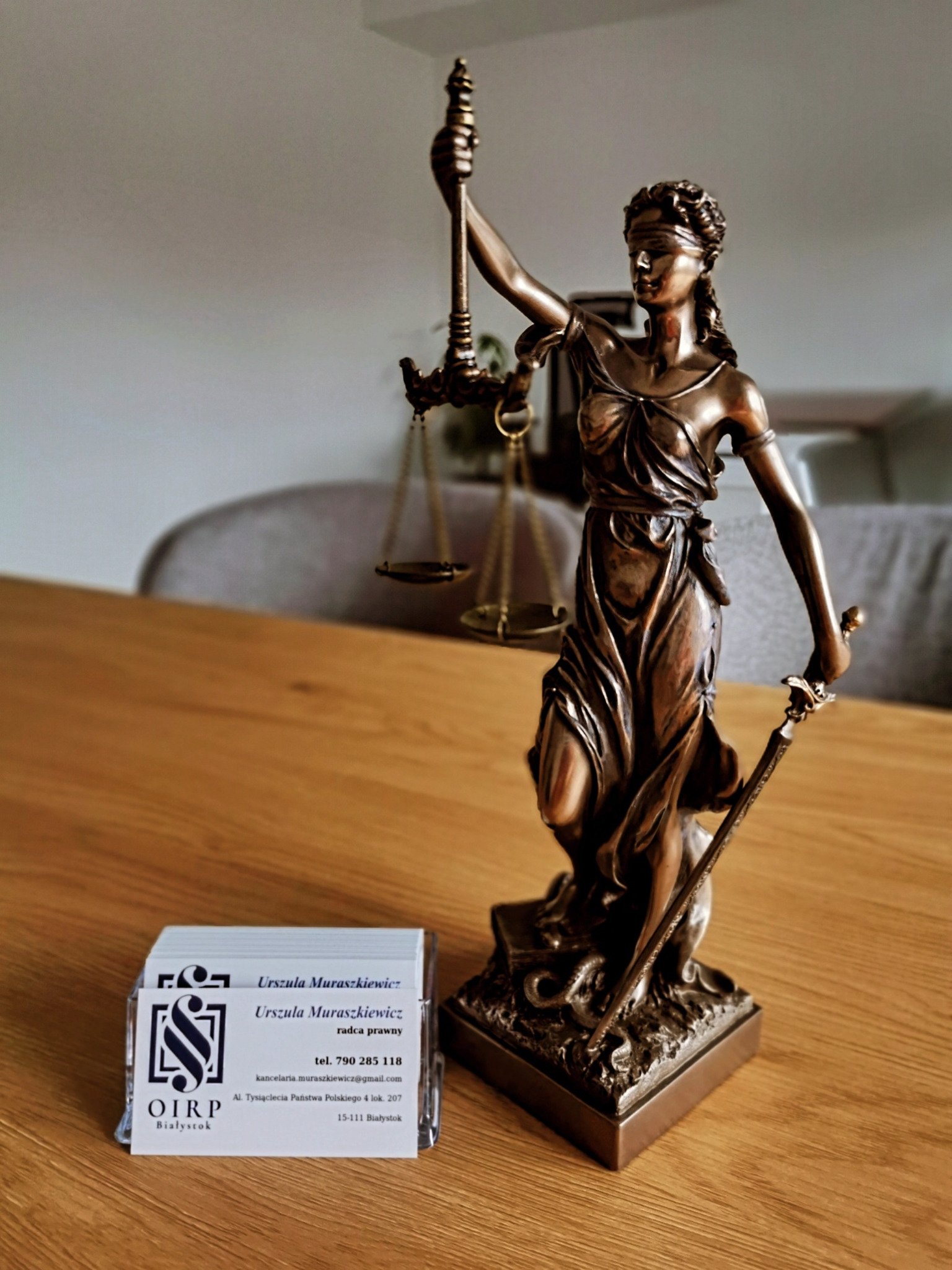 Statuetka Temidy z wagą i mieczem, wizytówki radcy prawnego Urszuli Muraszkiewicz na drewnianym biurku. Symbol sprawiedliwości w kancelarii.