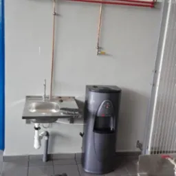 Szary dystrybutor wody z kranikiem i przyciskami obok metalowej umywalki, widoczne rury miedziane i czerwone na ścianie w pomieszczeniu o szarych płytkach.