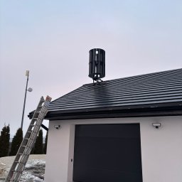 Montaż pionowej turbiny wiatrowej na dachu garażu. Aluminiowa drabina oparta o ścianę. W tle słup z anteną i ośnieżony teren.