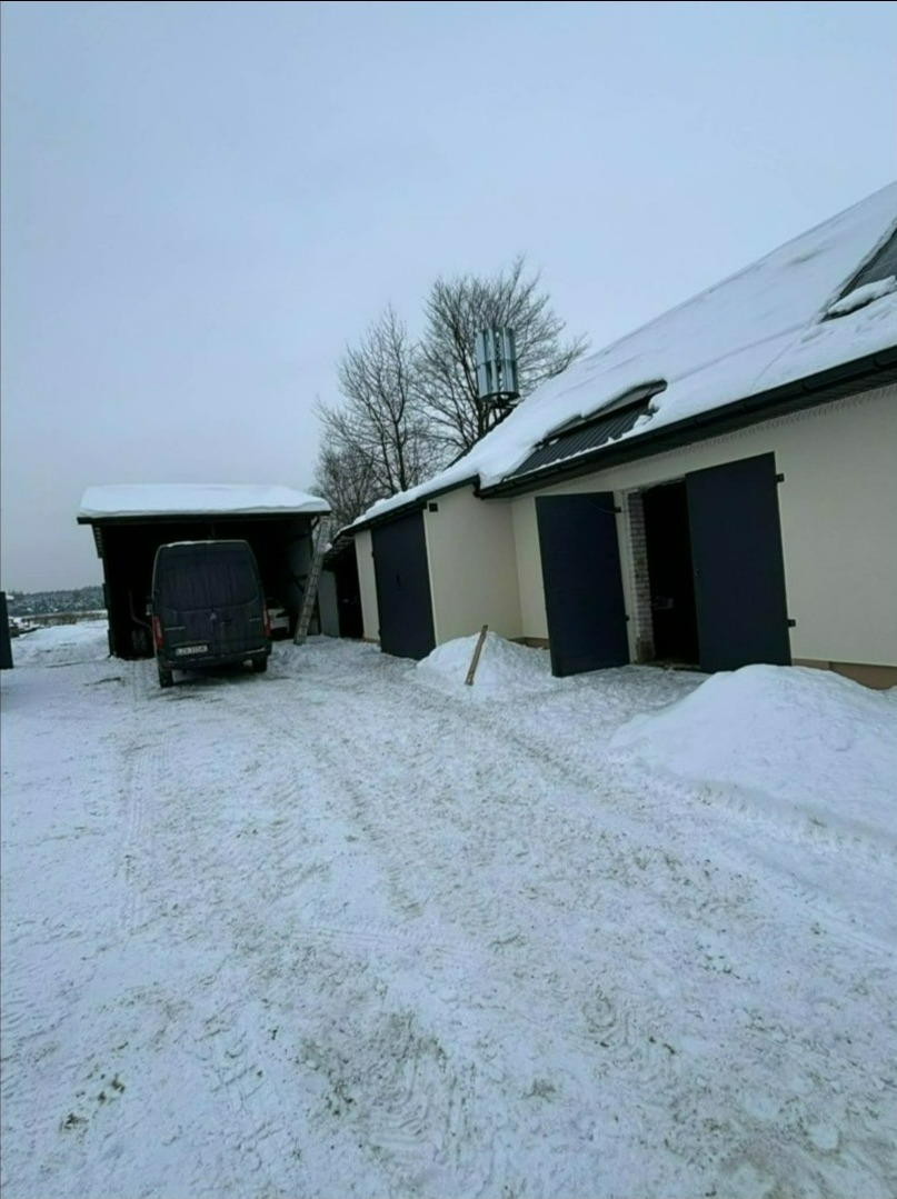 Zimowy krajobraz z budynkiem gospodarczym, na którego dachu widoczna turbina wiatrowa. Śnieg pokrywa teren i dachy budynków. Na pierwszym planie ślady opon na śniegu.