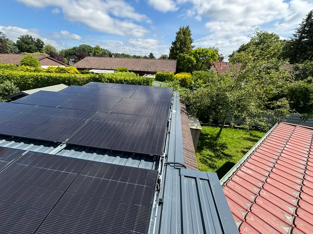 Czarny panel fotowoltaiczny na szarym dachu blaszany, widok z góry na tle zieleni i błękitnego nieba z chmurami. Czerwony dach sąsiedniego budynku w prawym dolnym rogu.