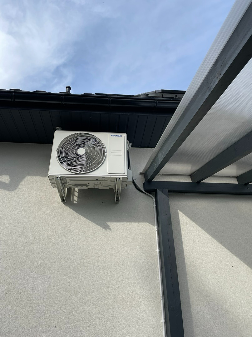 Biały klimatyzator Hyundai zamontowany na jasnej ścianie budynku, widoczne rury odprowadzające, zadaszenie w tle, ujęcie z dołu.