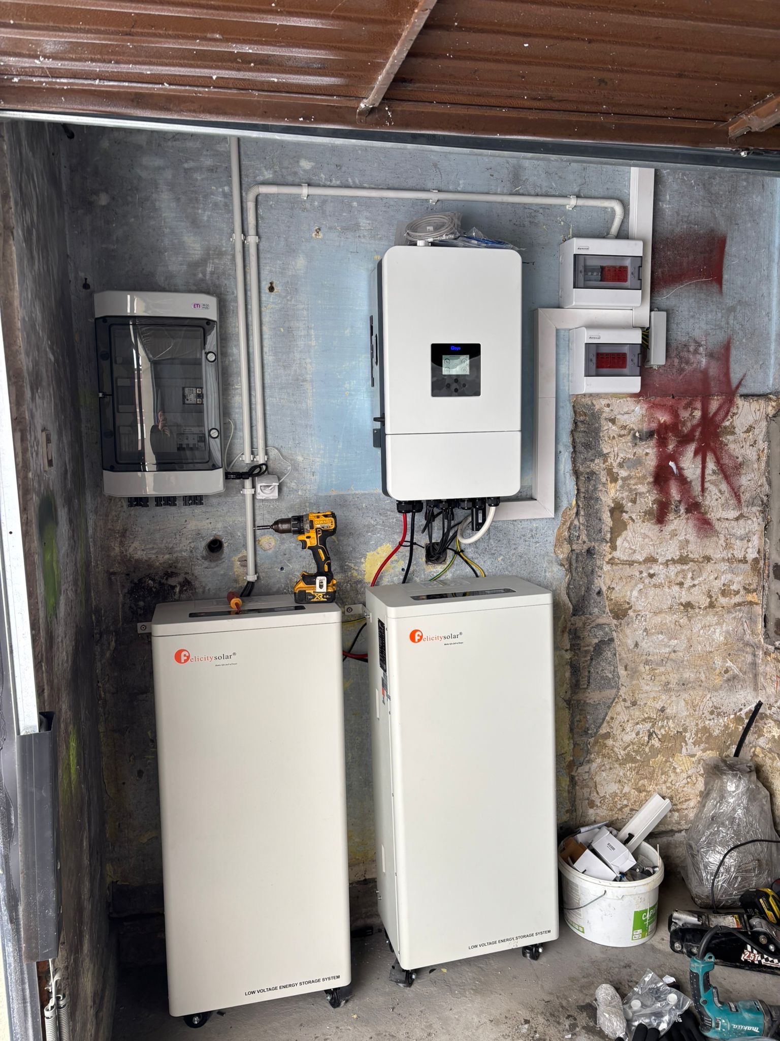 Instalacja magazynu energii Felicity Solar z inwerterem na tle surowej ściany. Widoczne przewody, skrzynki elektryczne i narzędzia. Montaż w garażu lub pomieszczeniu gospodarczym.