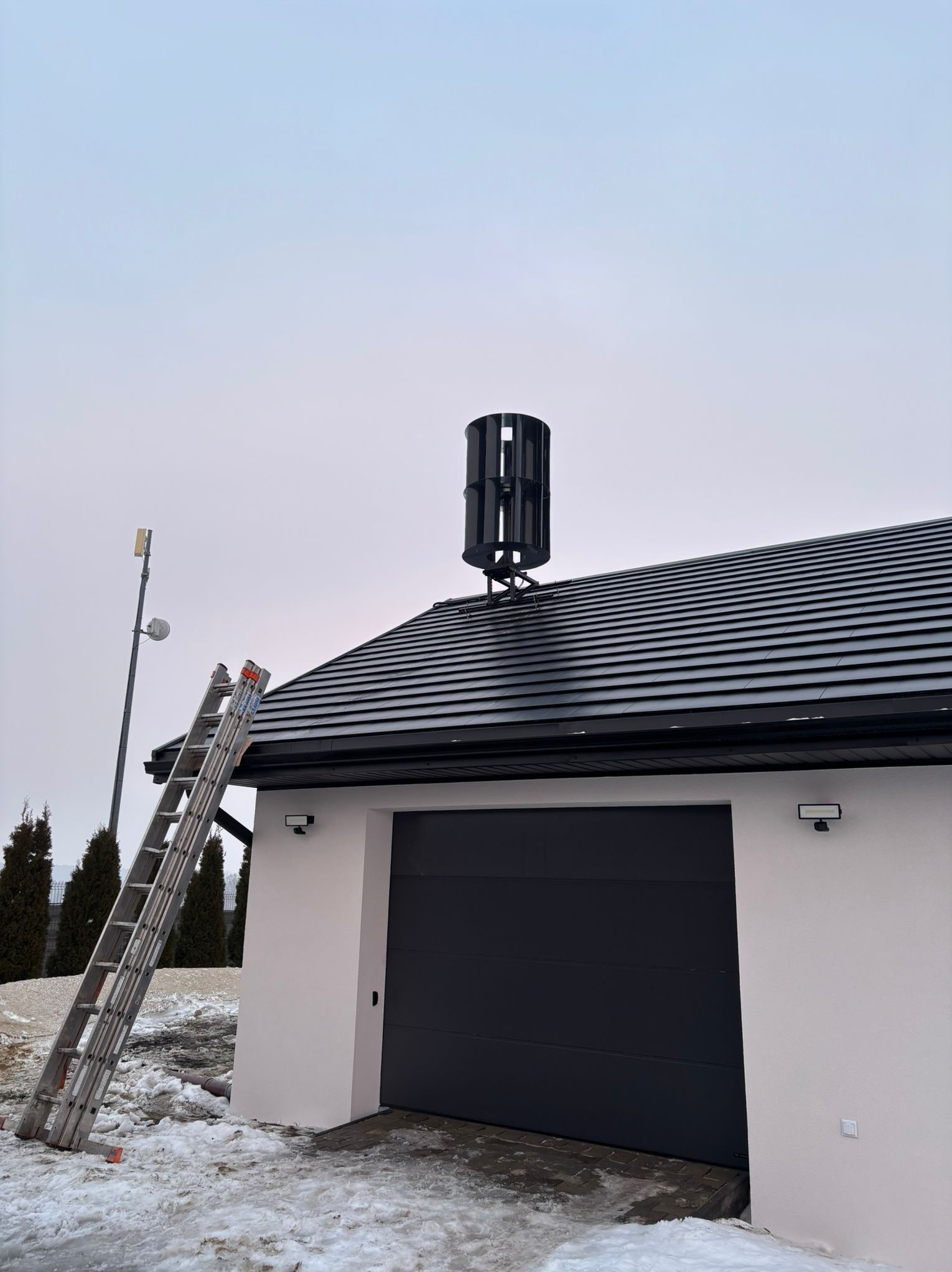 Montaż pionowej turbiny wiatrowej na dachu garażu. Aluminiowa drabina oparta o ścianę. W tle słup z anteną i ośnieżony teren.