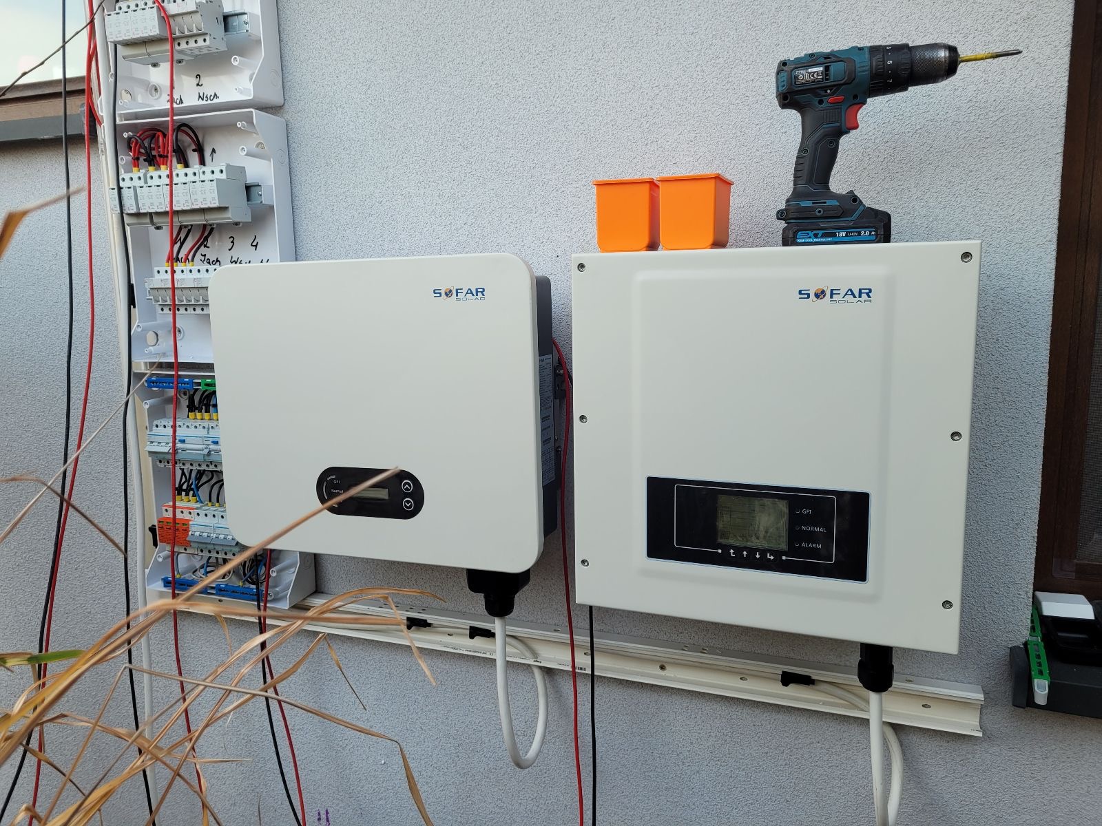 Dwa białe inwertery SOFAR SOLAR zamontowane na ścianie obok skrzynki elektrycznej. Na jednym z nich leżą pomarańczowe pojemniki i wiertarka.