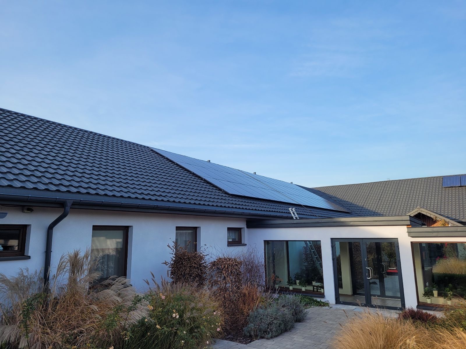 Biały dom z ciemną dachówką i panelami słonecznymi na dachu. Aluminiowa drabina oparta o dach. Ogród z ozdobnymi trawami na pierwszym planie.