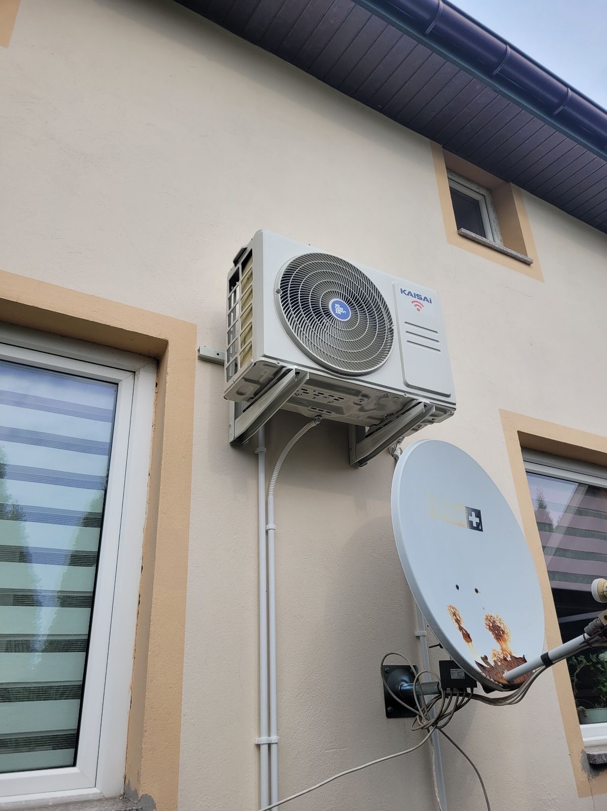 Biały klimatyzator Kaisai zamontowany na elewacji budynku, obok okna i anteny satelitarnej. Widoczne rury instalacyjne i uchwyty montażowe.