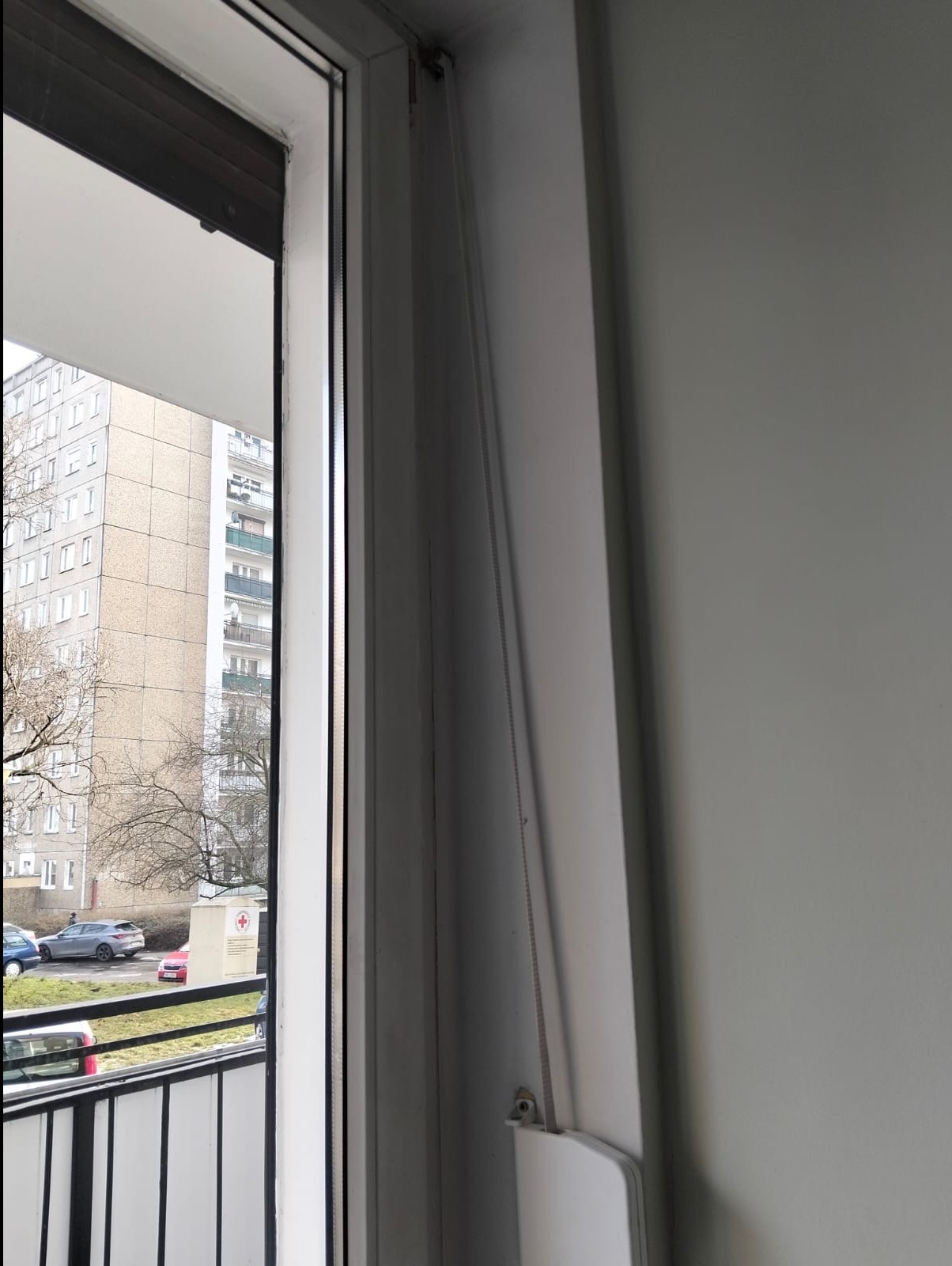 Fragment okna z częściowo opuszczoną, białą roletą wewnętrzną. Widok na blok mieszkalny i drzewa za oknem. Szara ściana w tle.