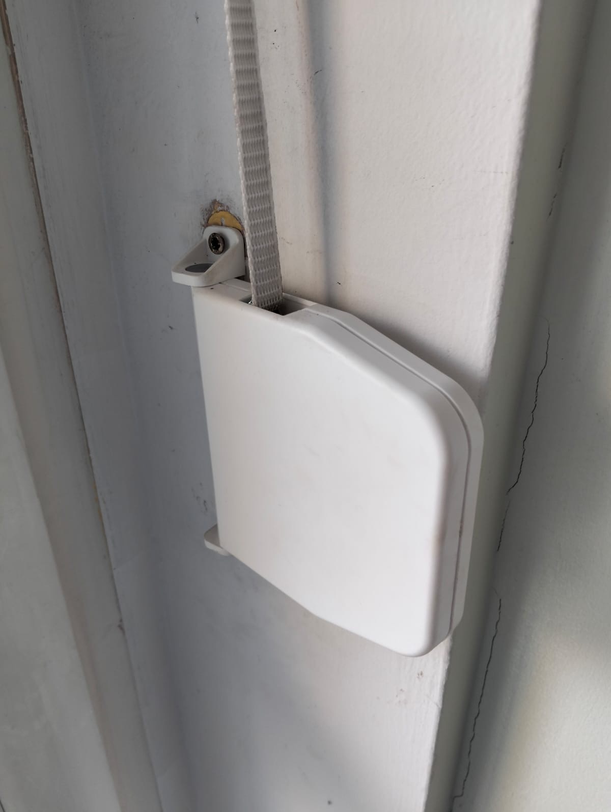 Biały mechanizm zwijający rolety przymocowany do ściany, widoczny pasek. Ściana z delikatnymi pęknięciami. Ujęcie z dołu, bliskie.