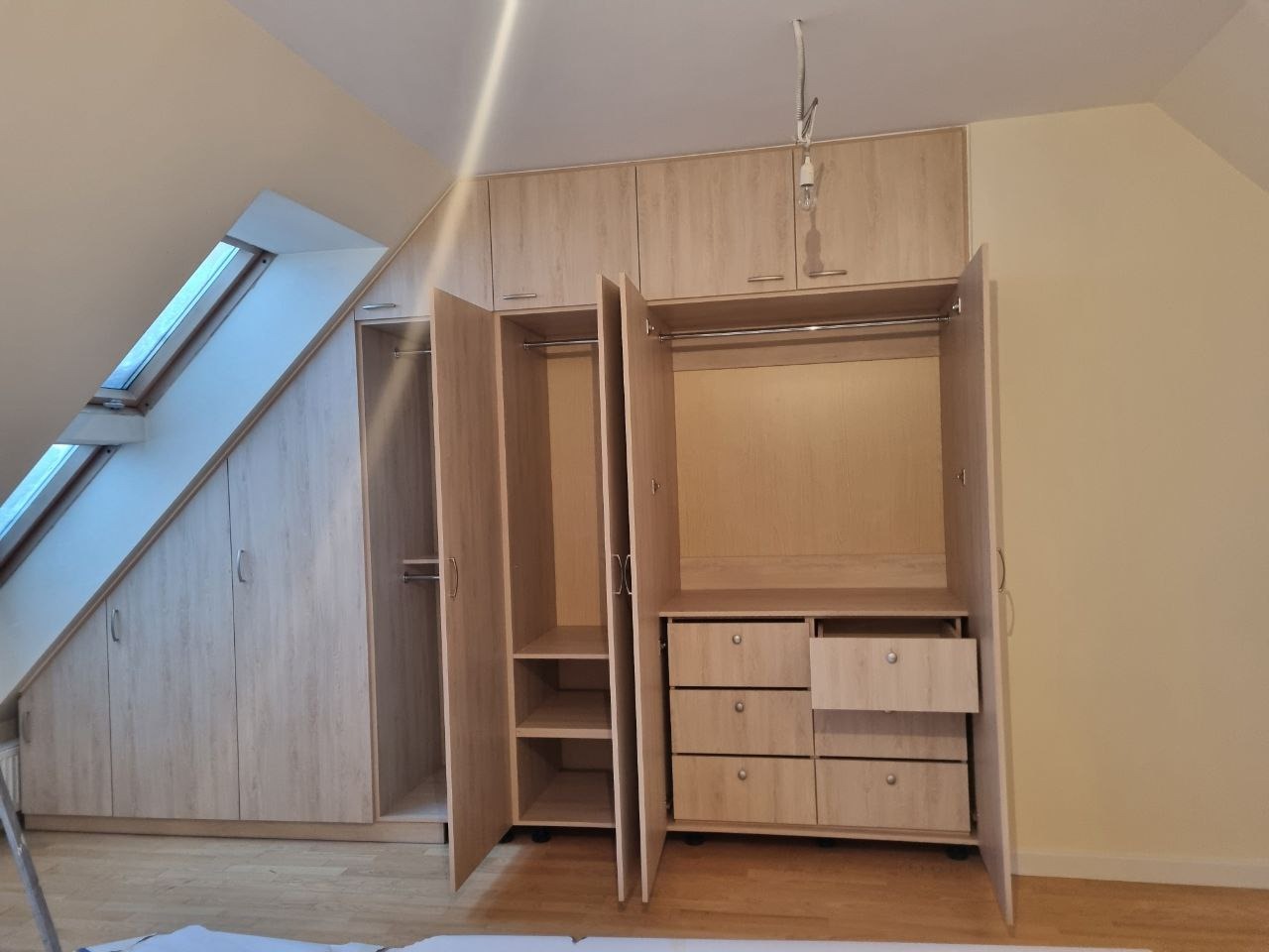 Jasna, drewnopodobna szafa wnękowa na poddaszu ze skosem, z otwartymi drzwiami ukazującymi półki i szuflady. Widoczne okno dachowe i oświetlenie sufitowe.