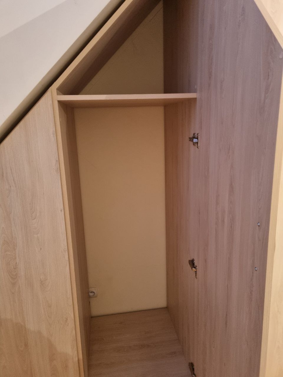 Otwarta szafa wnękowa z półką, wykonana z płyty imitującej drewno, dopasowana do skosu dachu. Widoczne zawiasy i gniazdko elektryczne wewnątrz.