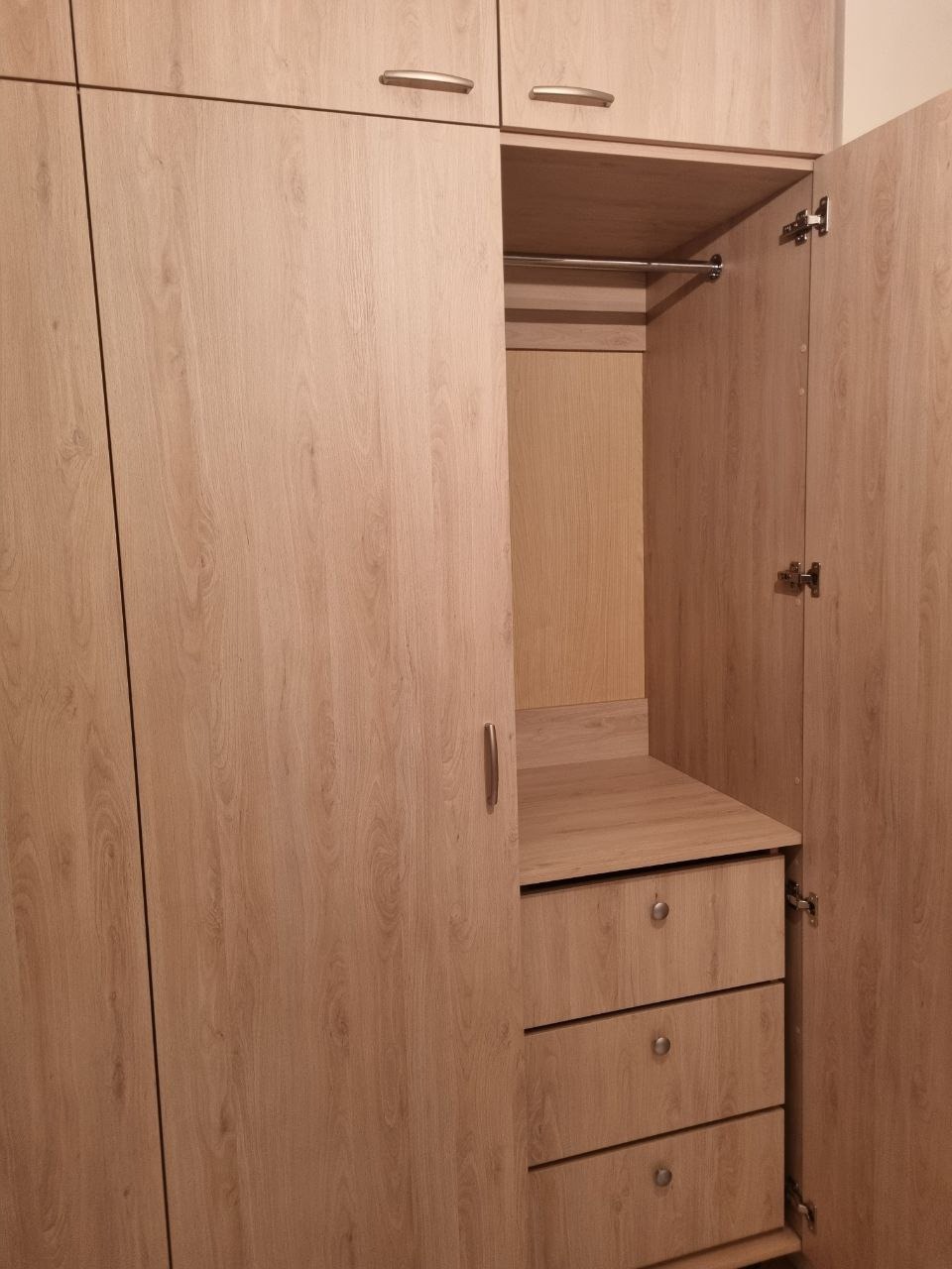 Jasna szafa z otwartymi drzwiami, ukazująca półki, drążek i szuflady. Widoczna faktura drewna i metalowe okucia. Ujęcie z bliska, wnętrze szafy dobrze oświetlone.