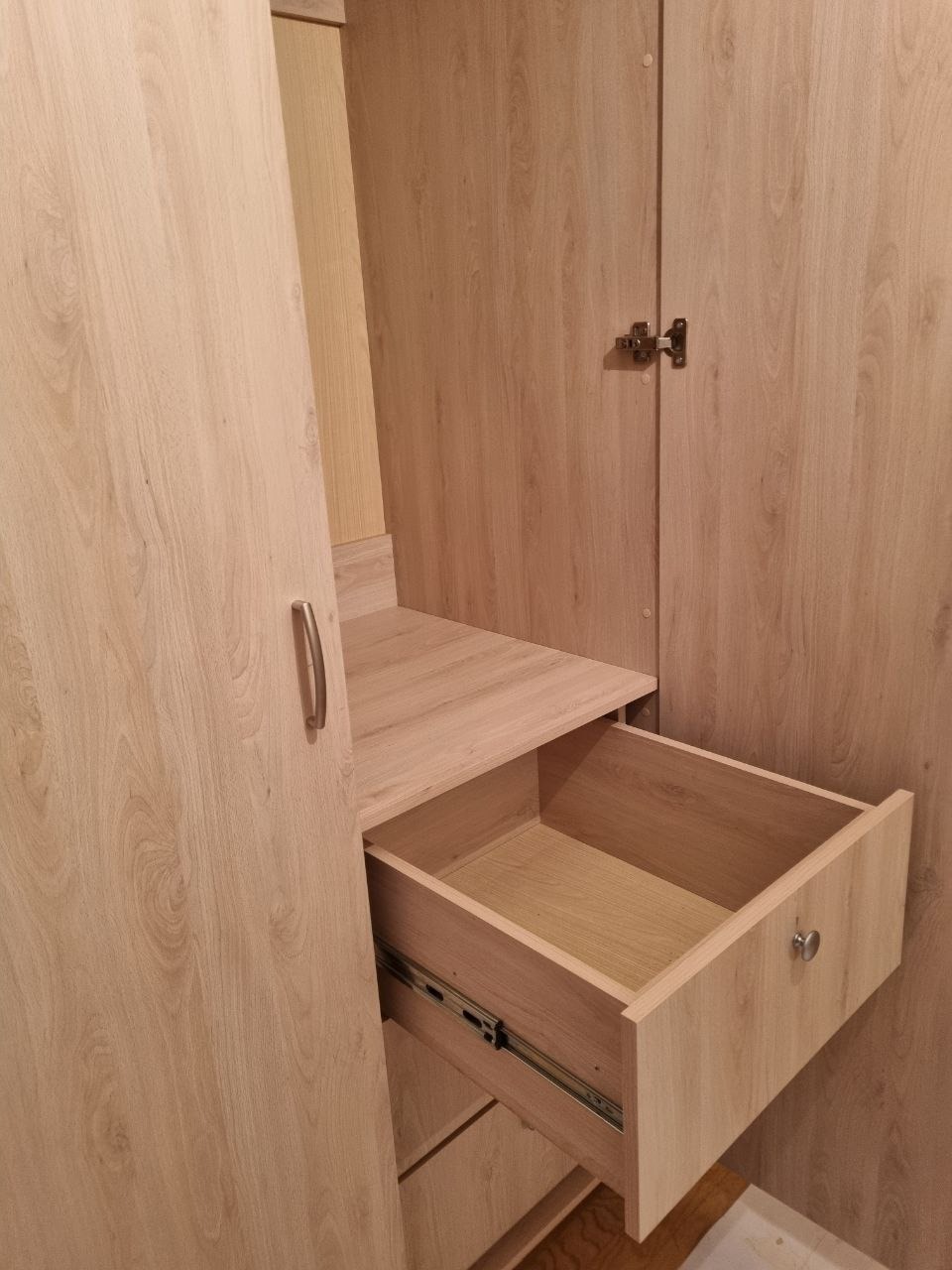 Jasna szafa z otwartą szufladą, widoczne prowadnice i półka. Drzwiczki szafy z metalowym uchwytem i zawiasami. Teksturowane drewno.