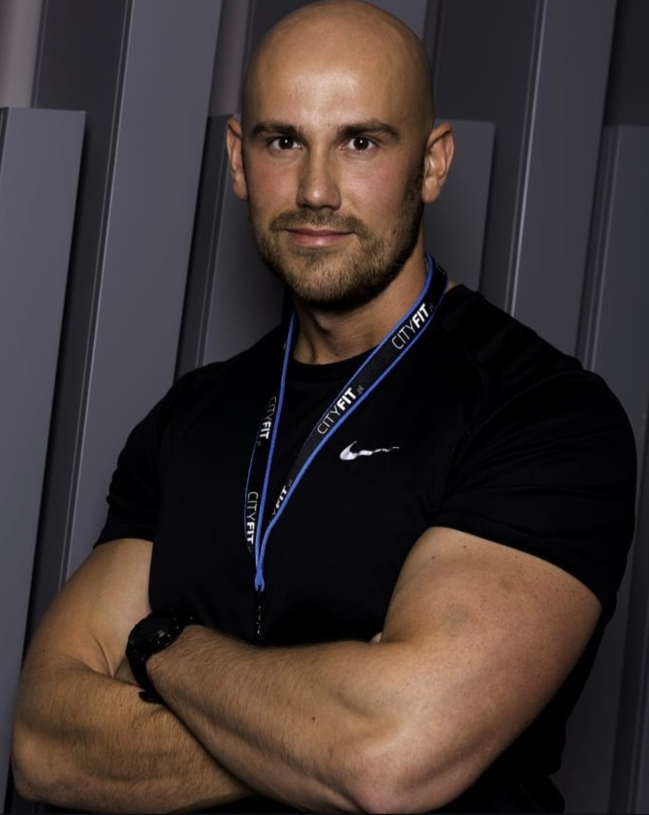 Portret mężczyzny z krótkimi włosami, w czarnej koszulce i identyfikatorem CityFit, z założonymi rękami na tle szarej ściany z pionowymi panelami.