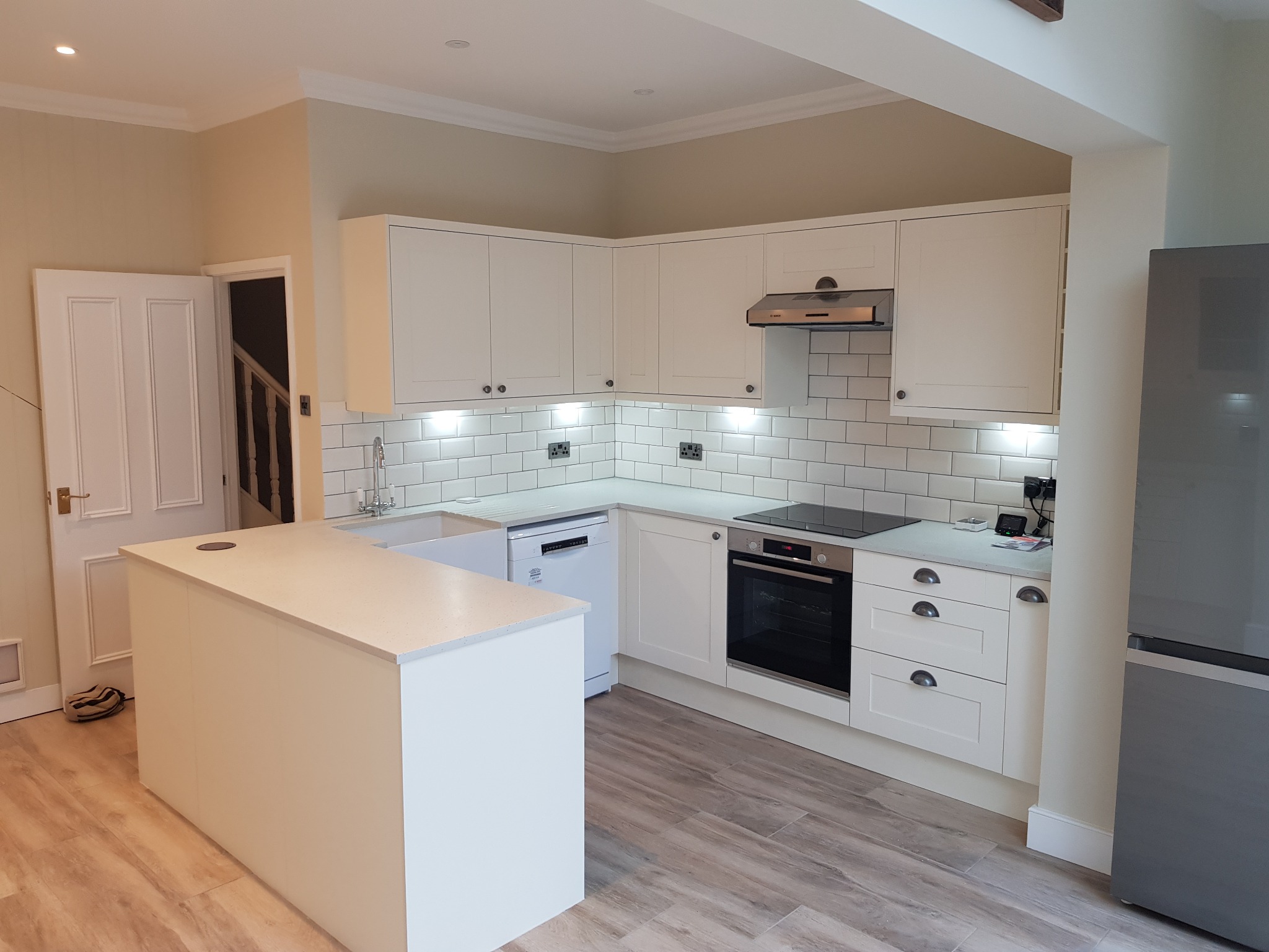 Jasna kuchnia po remoncie: białe szafki, płytki metro, drewnopodobna podłoga i wyspa kuchenna z blatem w kolorze jasnego kamienia.