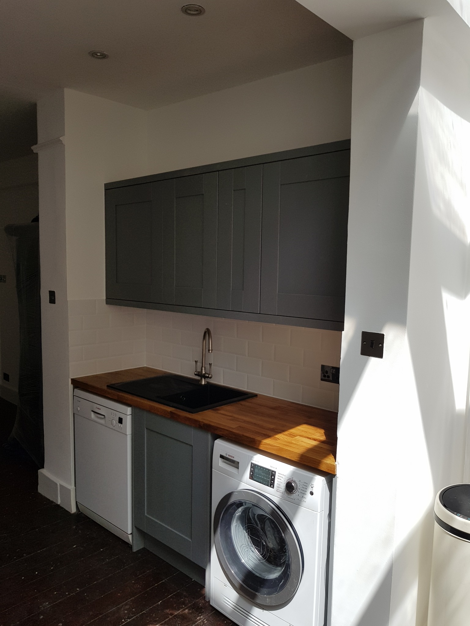Szare szafki kuchenne nad blatem z drewnianym wykończeniem, zlew, białe płytki na ścianie, pralka i zmywarka w jasnym pomieszczeniu z ciemną podłogą.
