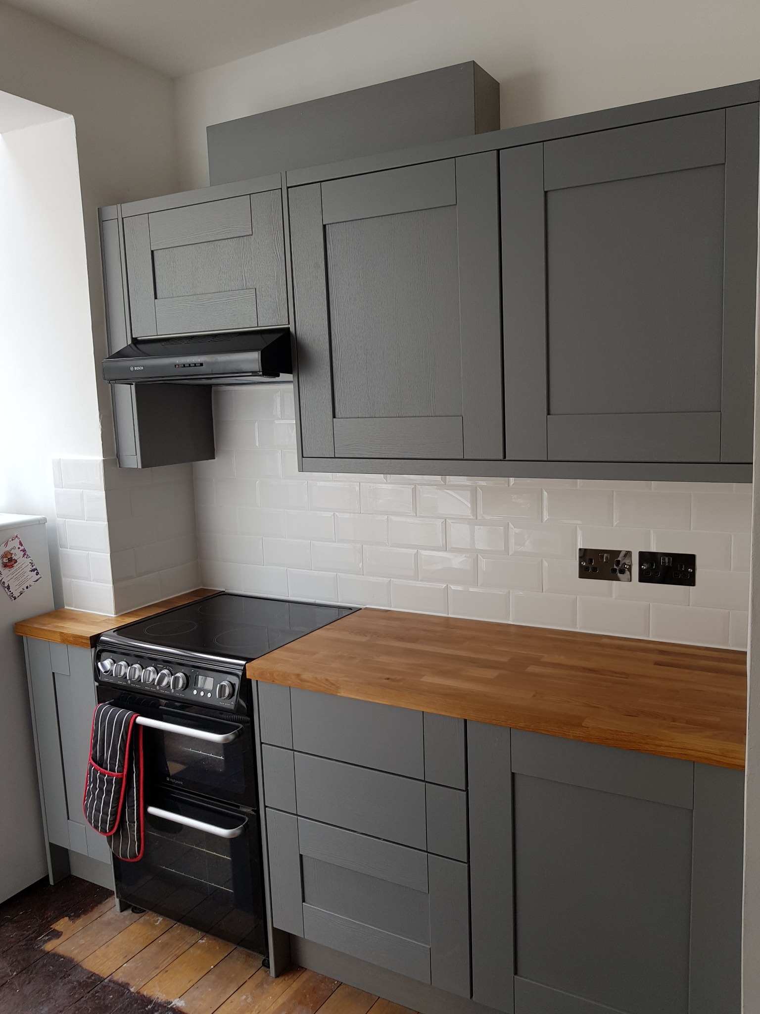 Szare szafki kuchenne z drewnianym blatem i białymi płytkami w Hecznarowicach. Widoczny okap i piekarnik. Klasyczny styl z nowoczesnymi akcentami.