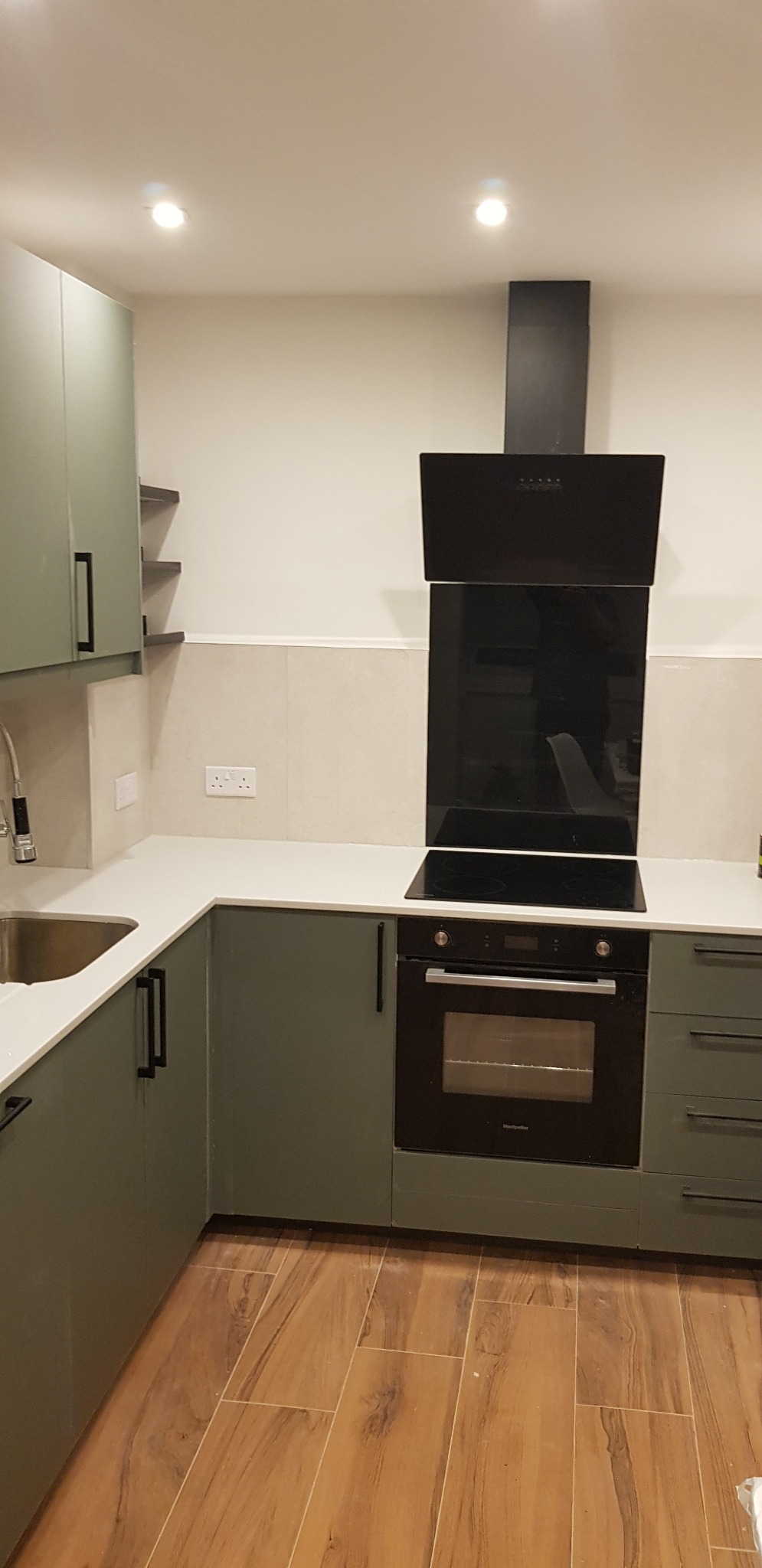 Nowoczesna kuchnia w odcieniach zieleni z czarnymi akcentami: okap, piekarnik i uchwyty. Jasny blat i drewnopodobna podłoga tworzą spójną całość.