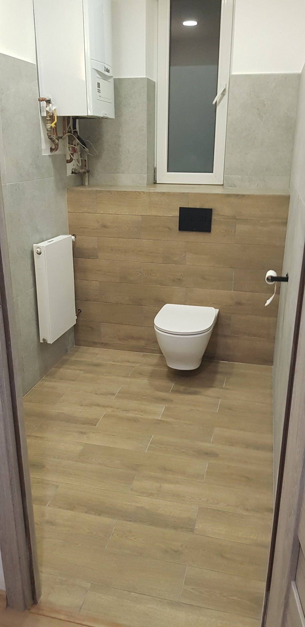 Wykończona łazienka z drewnopodobnymi płytkami na podłodze i ścianie, wiszącą toaletą i grzejnikiem. Widoczny piec gazowy i okno.