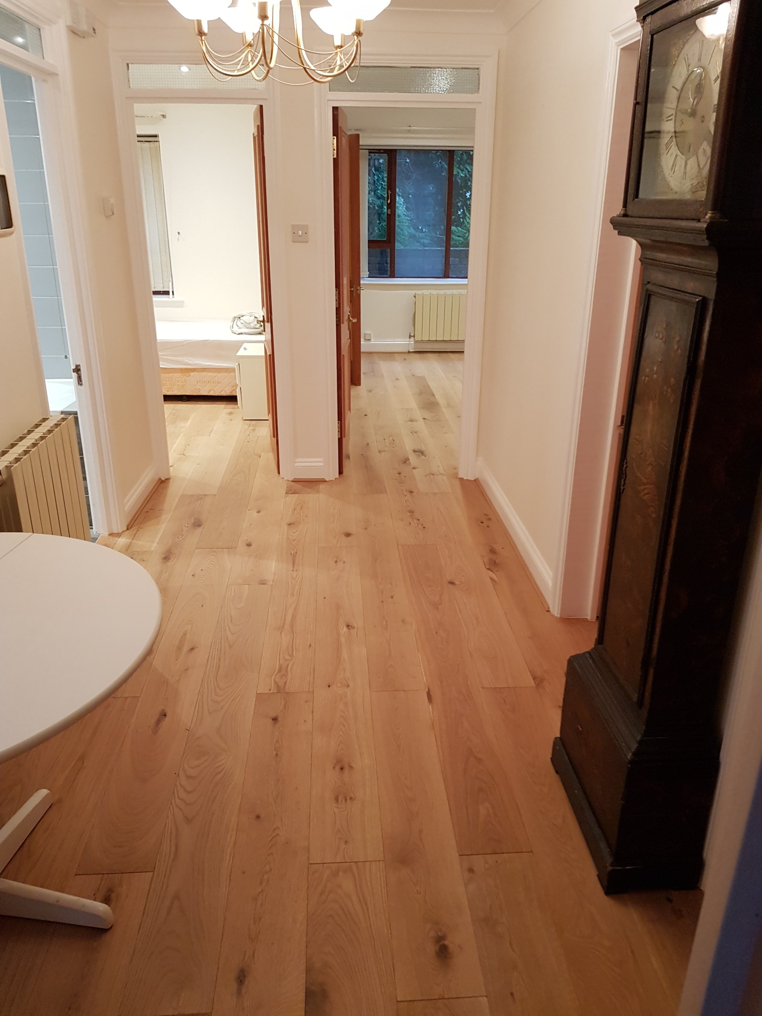 Jasne panele podłogowe w korytarzu z widokiem na pokoje, białe ściany i drzwi. W tle widoczny zegar stojący i fragment okrągłego stołu.