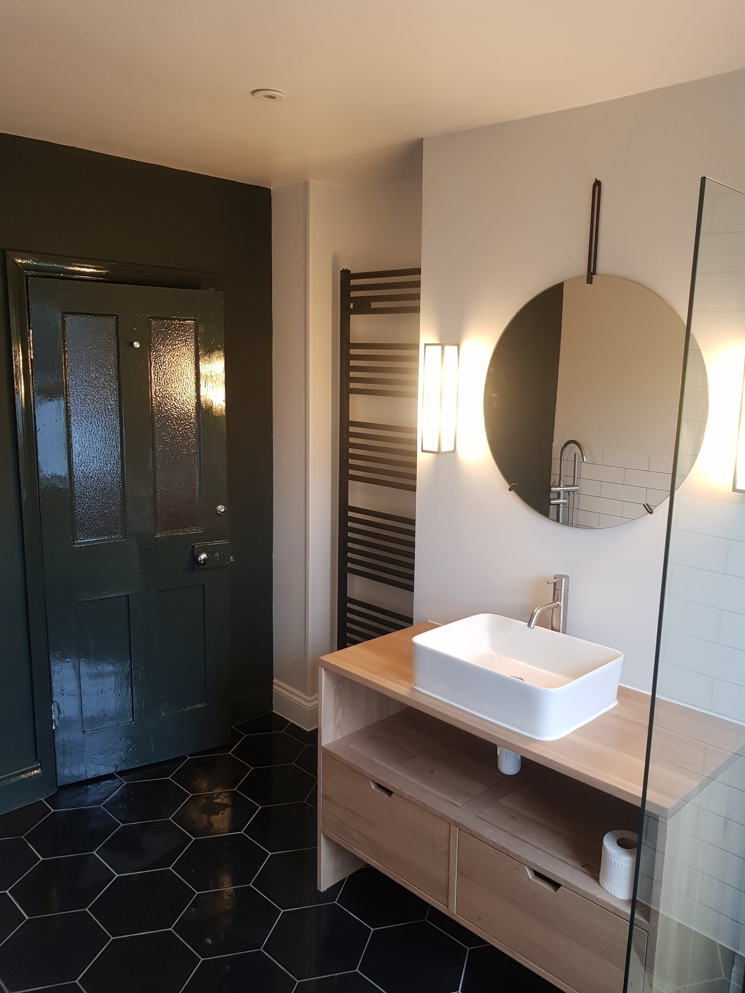 Nowoczesna łazienka z czarną, heksagonalną podłogą, drewnianą szafką pod umywalkę i okrągłym lustrem z podświetleniem. Ciemnozielone drzwi dodają elegancji.
