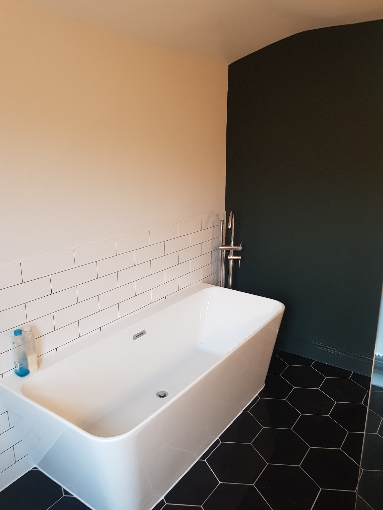 Nowoczesna łazienka z białą wanną wolnostojącą, czarną heksagonalną podłogą i ścianą w kolorze butelkowej zieleni. Minimalistyczny design, subtelne oświetlenie.