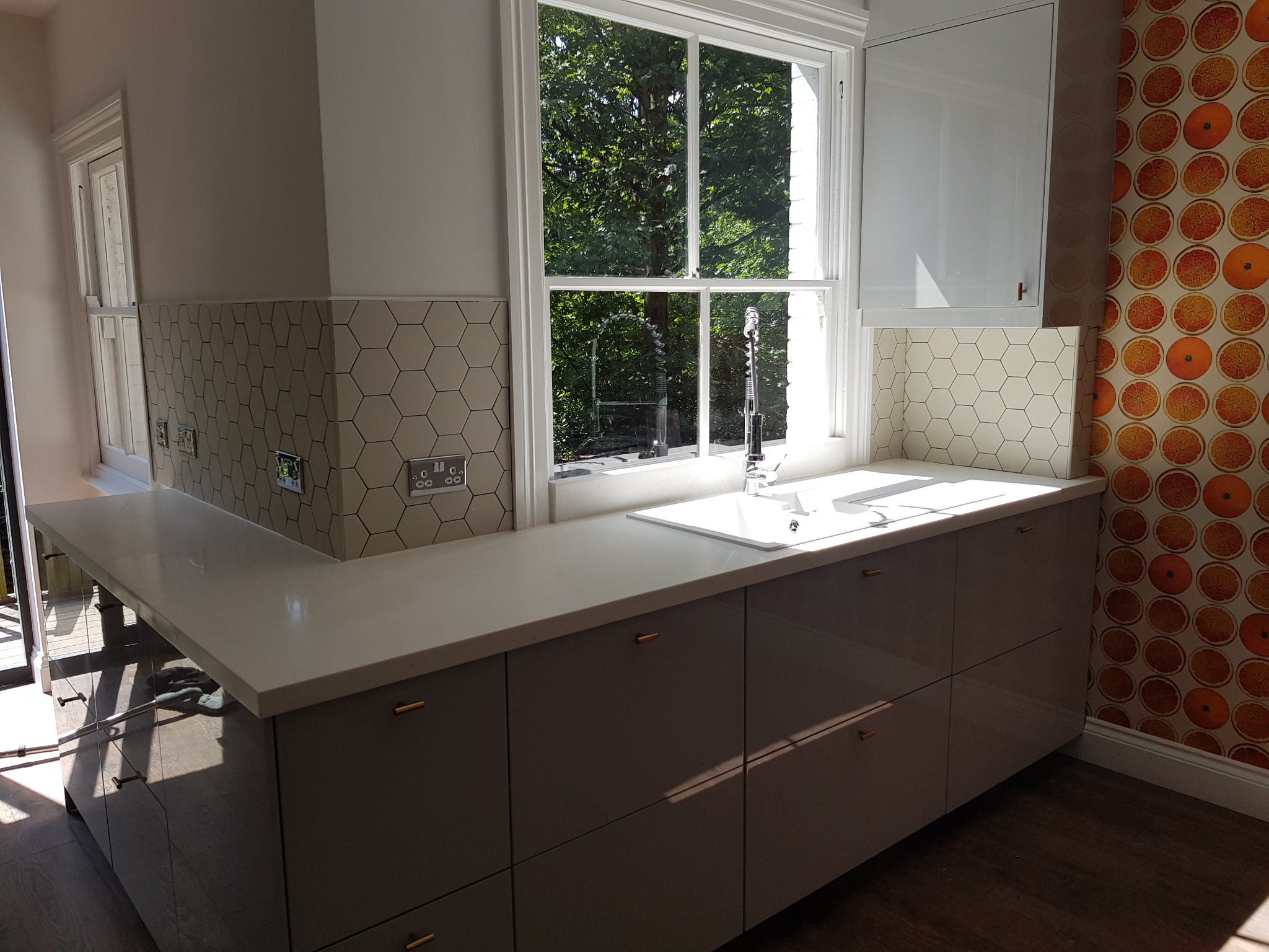 Jasna kuchnia po remoncie: szare szafki z złotymi uchwytami, biały blat, heksagonalne płytki nad blatem i okno z widokiem na zieleń. Tapeta w pomarańcze dodaje koloru.