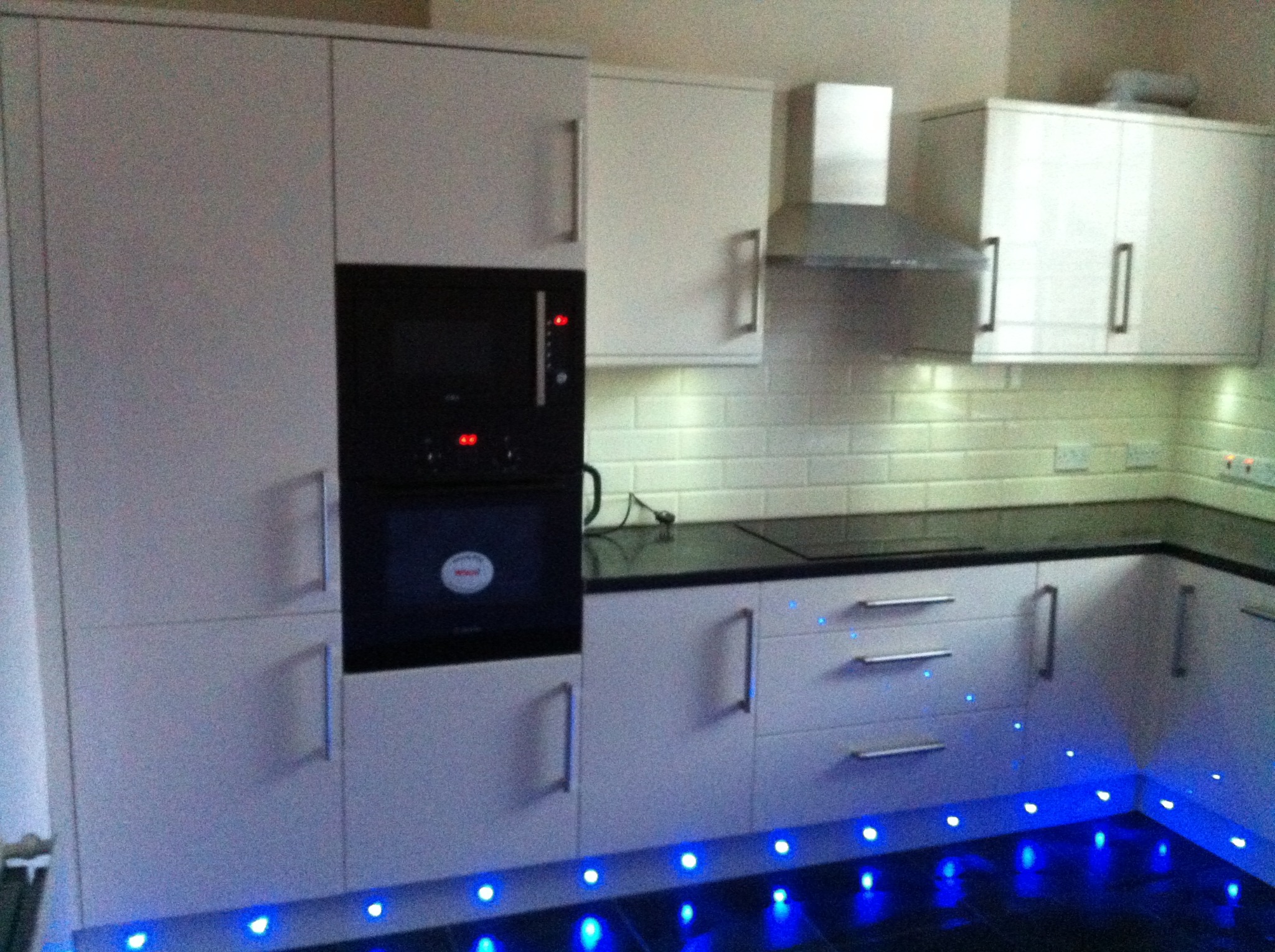 Biała, nowoczesna zabudowa kuchenna z piekarnikiem, mikrofalówką i okapem. Podświetlenie LED pod szafkami dodaje unikalnego charakteru. Białe płytki na ścianie.