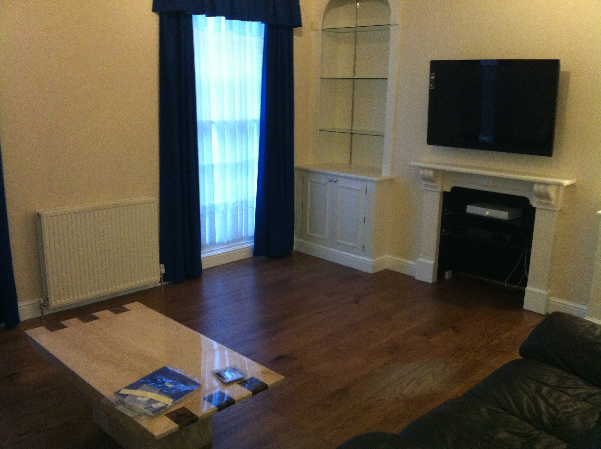 Salon po remoncie: drewniana podłoga, kremowe ściany, zabudowa z półkami, telewizor nad kominkiem. Nowoczesny stolik kawowy i zasłony w oknie.