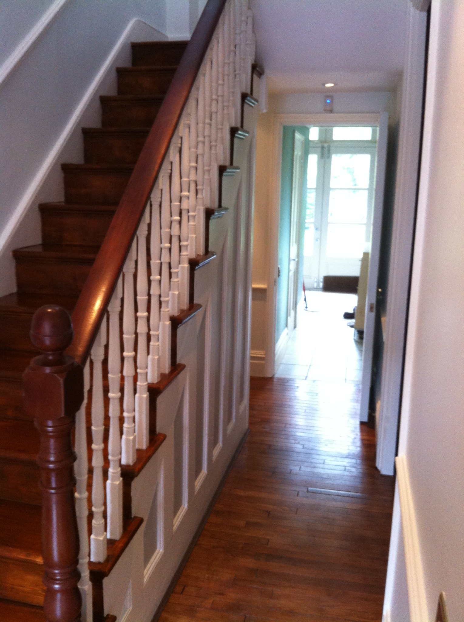 Drewniane schody z białą balustradą i panelami ściennymi w jasnym wnętrzu. Parkiet na podłodze, w tle widoczne drzwi i okno.