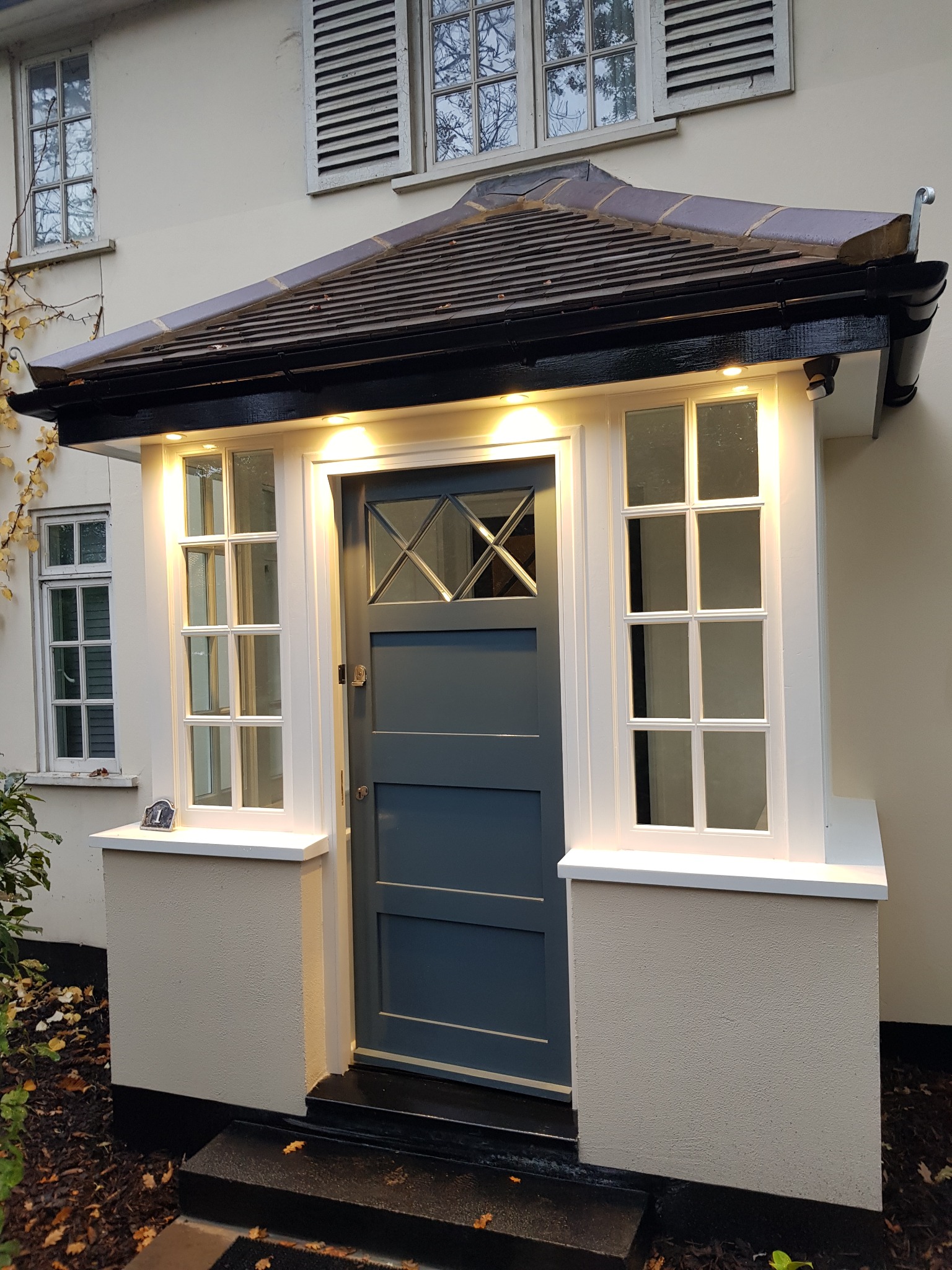 Odnowione wejście do domu z szarą stolarką drzwiową i białymi oknami po bokach, oświetlone ciepłym światłem. Widoczne detale wykończenia i jesienne liście.