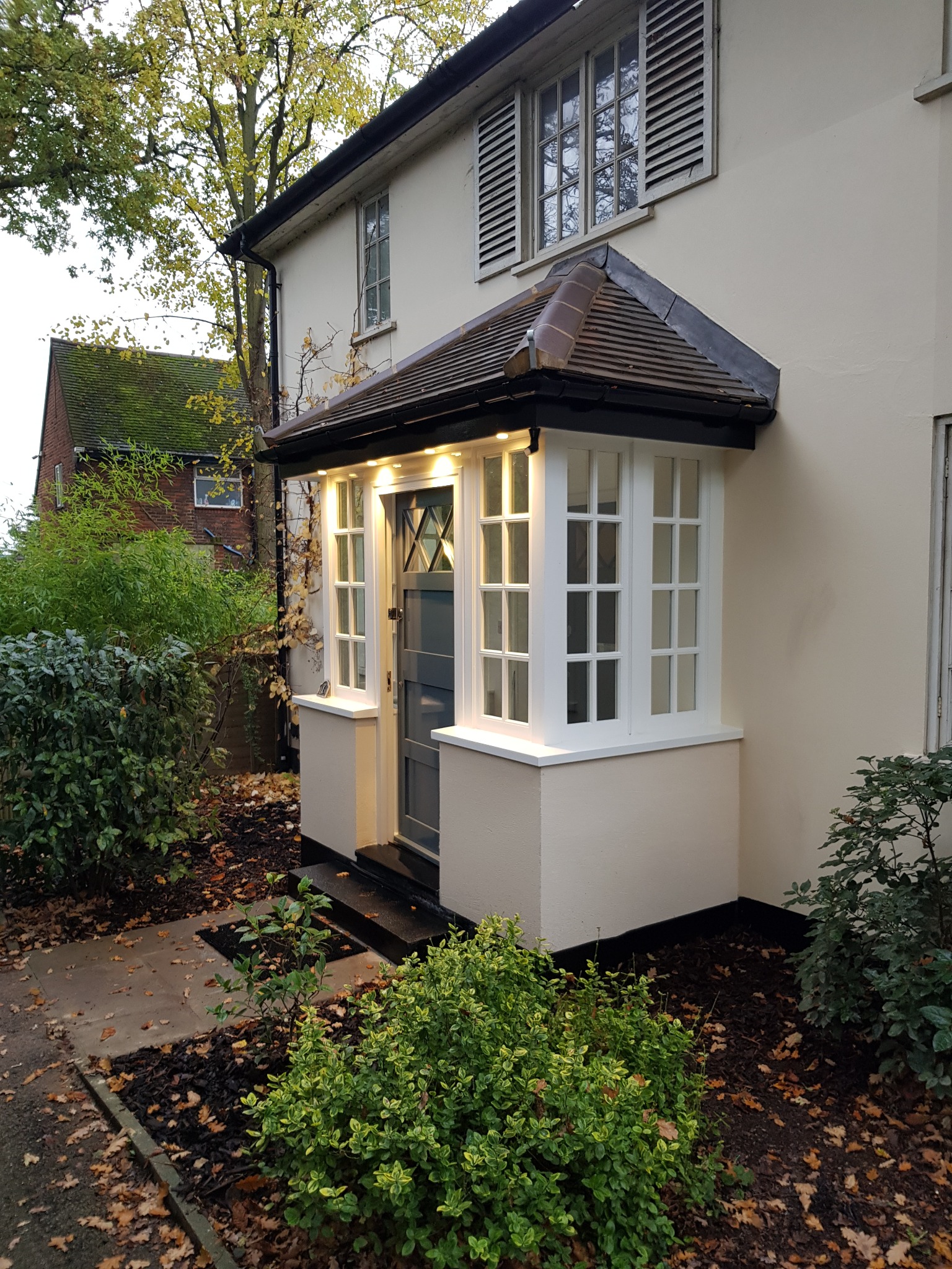 Odnowione wejście do domu z oświetlonym gankiem, białą stolarką okienną i szarą stolarką drzwiową. Elewacja w jasnym odcieniu beżu. Jesienne otoczenie z opadłymi liśćmi.