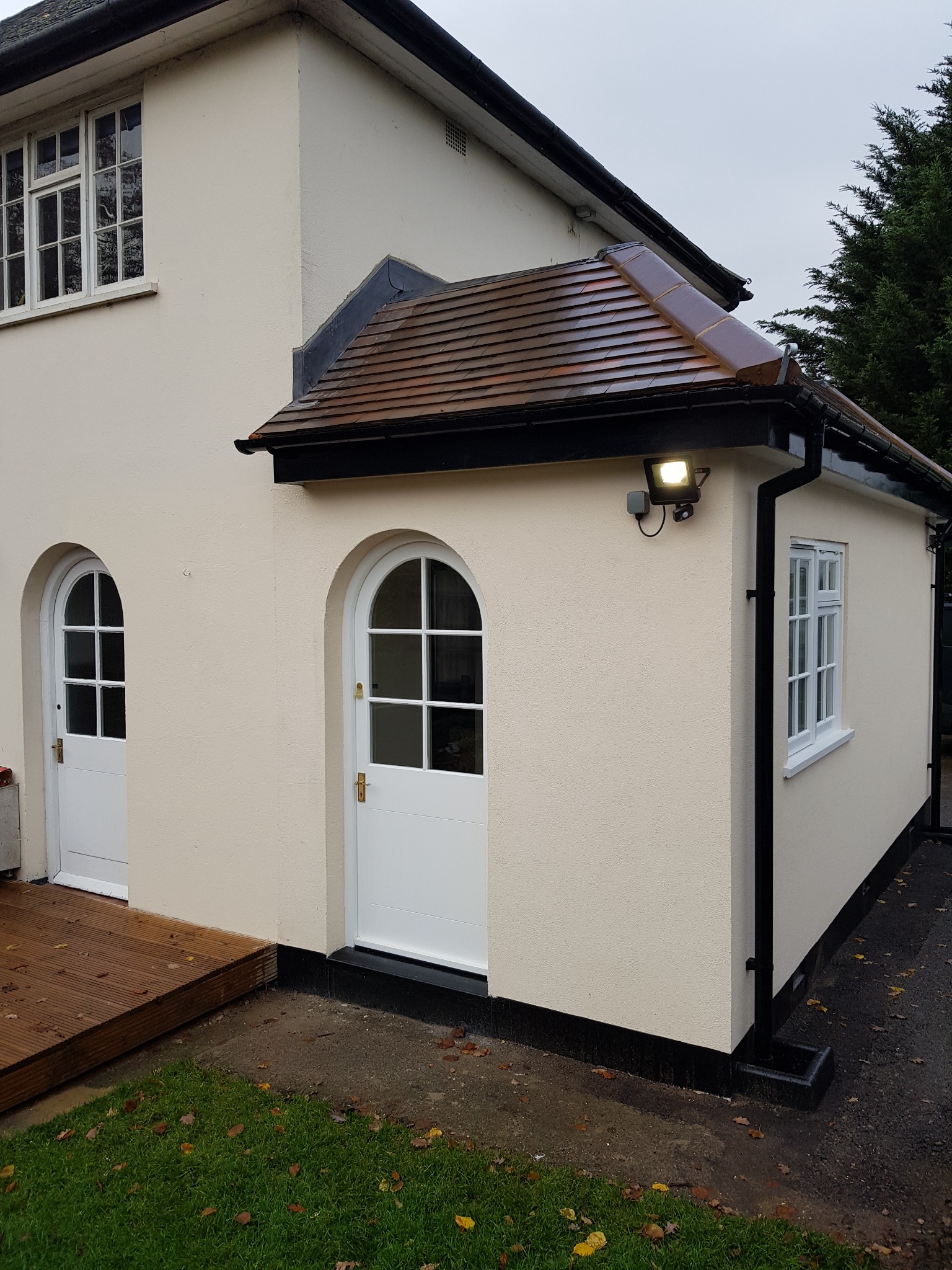 Elewacja budynku po remoncie z dwiema białymi, łukowatymi drzwiami i oknem. Widoczne brązowe dachówki i oświetlenie zewnętrzne. Świeżo pomalowana fasada w kolorze kremowym.