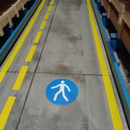 PREVENTO Serwis Techniczno - Porządkowy Paweł Skubiś - Oznakowanie magazynu: żółte linie wyznaczają ścieżkę dla pieszych, oznaczona niebieskim piktogramem. Metalowe regały z towarem po bokach. Betonowa posadzka.