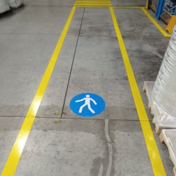 PREVENTO Serwis Techniczno - Porządkowy Paweł Skubiś - Oznakowanie poziome w hali magazynowej: żółte linie wyznaczają ścieżkę dla pieszych, z niebieskim symbolem pieszego na podłodze. Widoczne regały i zapakowane towary.