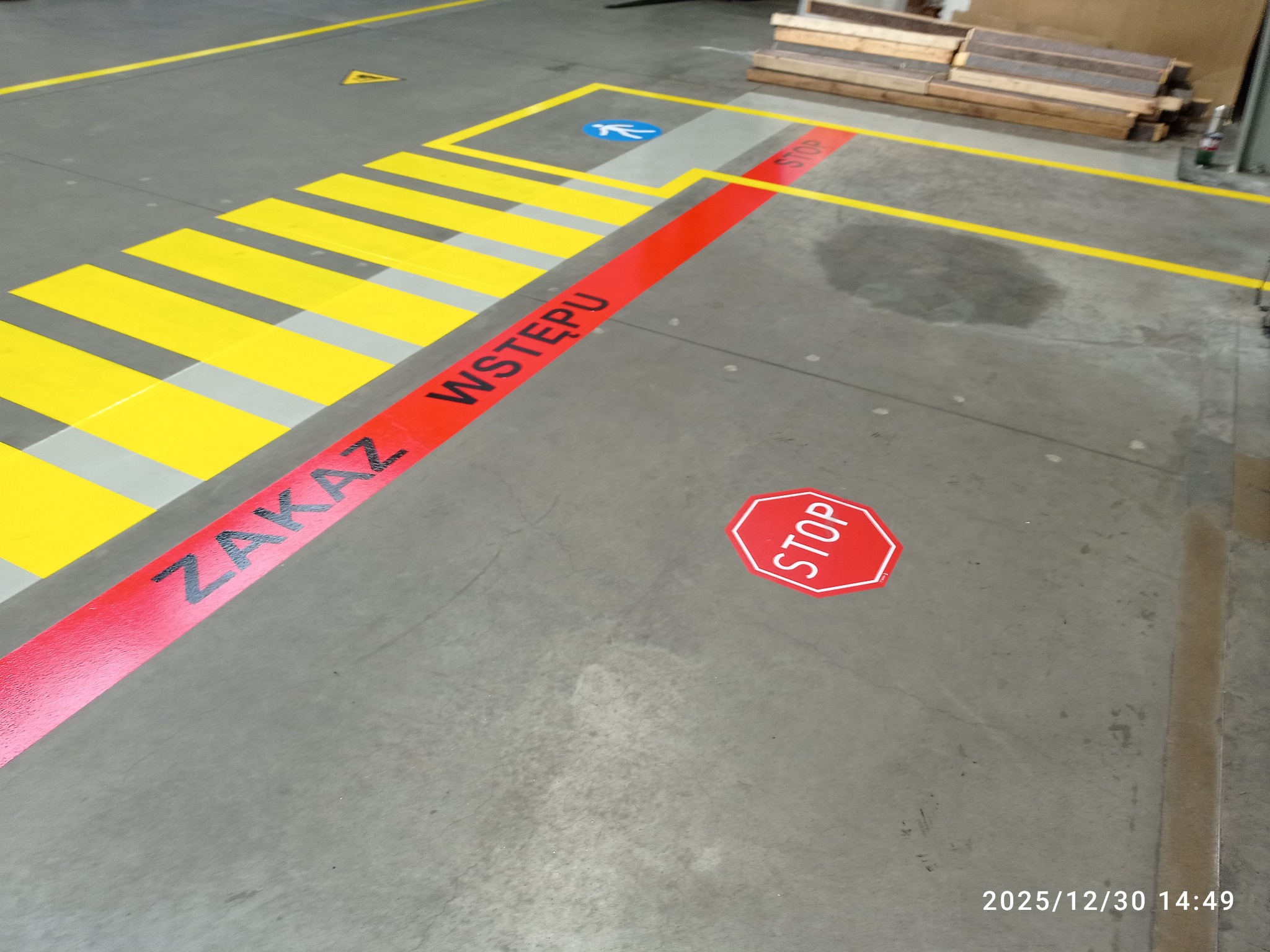 Oznakowanie poziome magazynu: żółto-szare pasy przejścia, czerwona linia z napisem 'Zakaz Wstępu' i znak 'STOP' na szarym betonie. Widoczne palety w tle.
