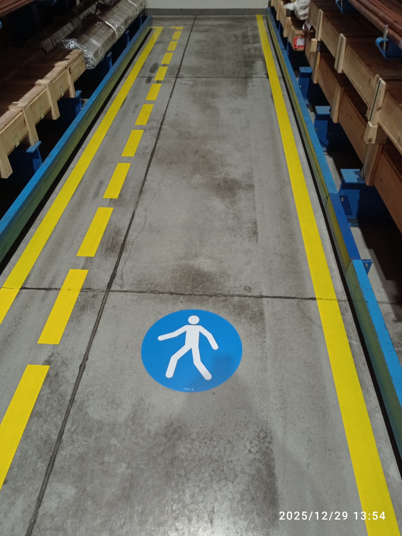 Oznakowanie magazynu: żółte linie wyznaczają ścieżkę dla pieszych, oznaczona niebieskim piktogramem. Metalowe regały z towarem po bokach. Betonowa posadzka.