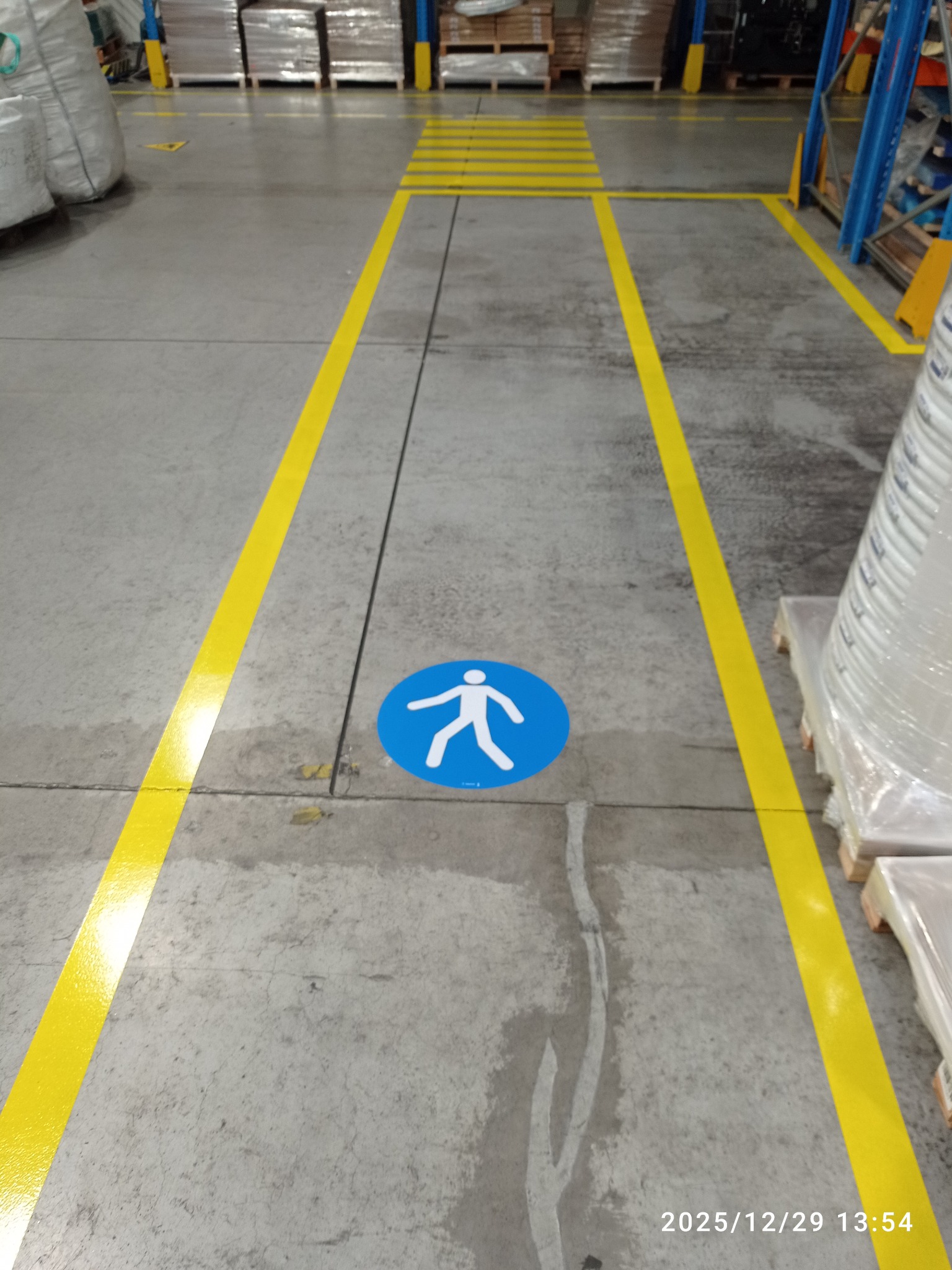 Oznakowanie poziome w hali magazynowej: żółte linie wyznaczają ścieżkę dla pieszych, z niebieskim symbolem pieszego na podłodze. Widoczne regały i zapakowane towary.