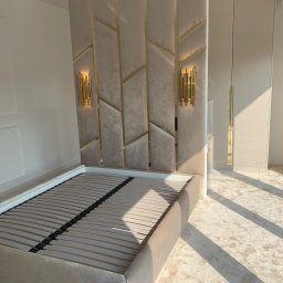 IPERFE - Beżowe łóżko tapicerowane z zagłówkiem w geometryczne złote wzory i lampkami, na tle białej ściany i szafy. Promienie słoneczne na dywanie.