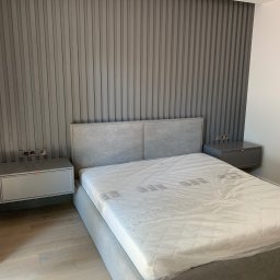IPERFE - Szara sypialnia z łóżkiem tapicerowanym i panelami ściennymi. Nocne szafki wiszące z gniazdkami elektrycznymi. Minimalistyczny design.
