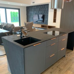 IPERFE - Nowoczesna wyspa kuchenna z czarnym blatem i szarymi szafkami w Radomiu. Minimalistyczny design, drewniana podłoga i heksagonalna ściana w tle.