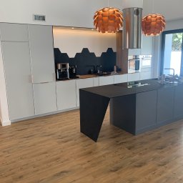 IPERFE - Nowoczesna kuchnia z szarymi szafkami, czarnym blatem wyspy i heksagonalnymi płytkami na ścianie. Nad wyspą wiszą dwie lampy w kształcie kuli i metalowy okap.