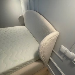 IPERFE - Beżowe łóżko z wełnianym zagłówkiem w minimalistycznej sypialni. Widoczny materac i fragment podłogi. Nad łóżkiem lampa. Neutralna kolorystyka.