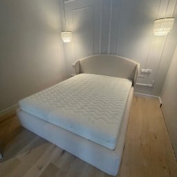 IPERFE - Eleganckie łóżko tapicerowane w jasnej tonacji, z wysokim wezgłowiem i materacem, w minimalistycznej sypialni z drewnianą podłogą i dekoracyjnymi lampami wiszącymi.