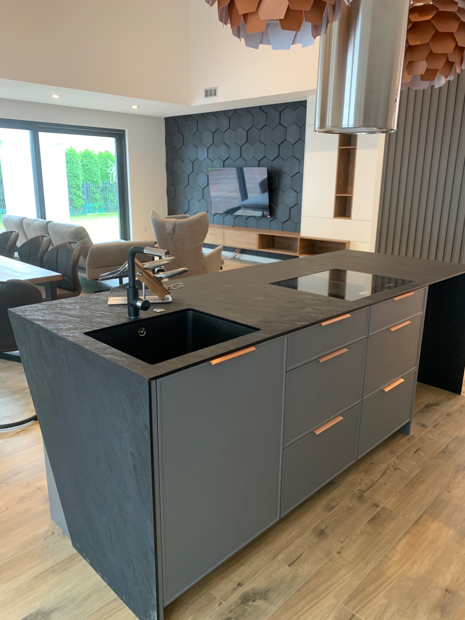 Nowoczesna wyspa kuchenna z czarnym blatem i szarymi szafkami w Radomiu. Minimalistyczny design, drewniana podłoga i heksagonalna ściana w tle.